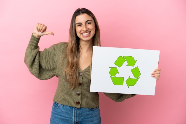 Młoda kobieta kaukaski na białym tle na różowym tle, trzymając plakietkę z ikoną recyklingu z dumnym gestem