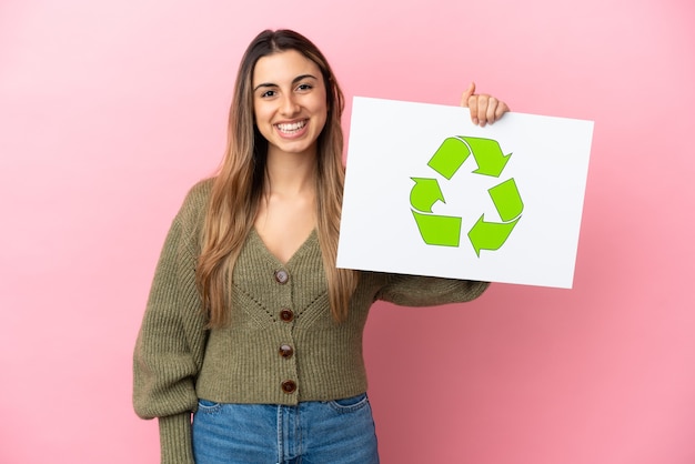 Młoda kobieta kaukaski na białym tle na różowym tle, trzymając plakietkę z ikoną recyklingu z happy wypowiedzi