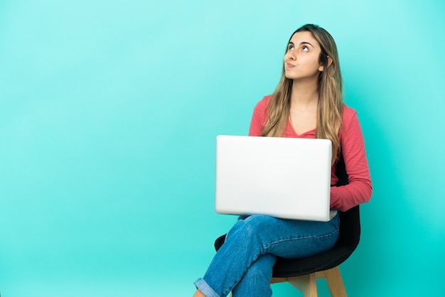 Młoda kobieta kaukaski siedzi na krześle z jej komputera na białym tle na niebieskiej ścianie i patrząc w górę
