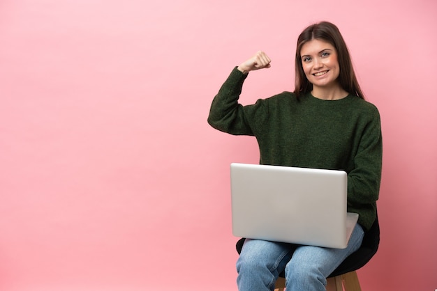 Młoda kobieta kaukaski siedzi na krześle z laptopem na białym tle na różowym tle robi silny gest