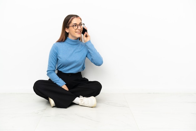 Młoda kobieta Kaukaski siedzi na podłodze, prowadząc rozmowę z telefonem komórkowym