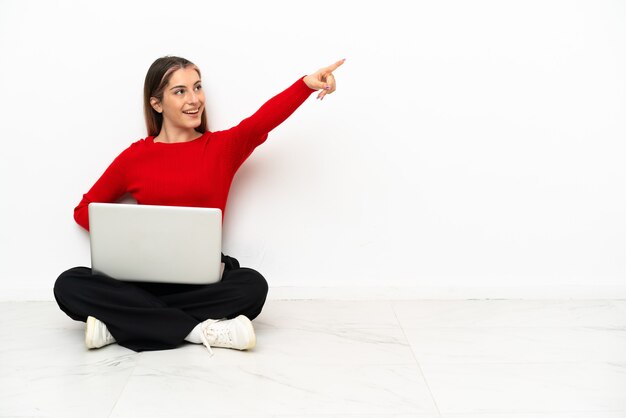 Młoda kobieta kaukaski z laptopem siedzi na podłodze, wskazując dalej