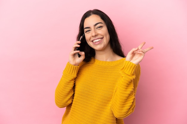 Młoda Kobieta Kaukaski Za Pomocą Telefonu Komórkowego Na Białym Tle Na Różowej ścianie, Uśmiechając Się I Pokazując Znak Zwycięstwa
