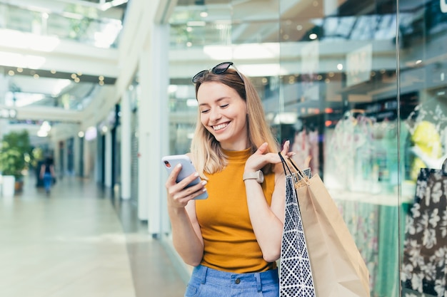 Młoda kobieta konsumentka w centrum handlowym przegląda czat i korzysta ze smartfona. kobieta stojąca z telefonem komórkowym w dłoniach w centrum handlowym. wnętrz. szczęśliwa dziewczyna z torby na prezenty robi zakupy