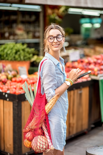Młoda kobieta korzystająca ze smartfona stojąca z torbą przed targiem spożywczym we Francji