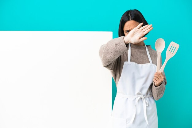 Młoda kobieta kucharz rasy mieszanej z dużą tabliczką odizolowaną na niebieskim tle, wykonując gest zatrzymania ręką, aby zatrzymać akt