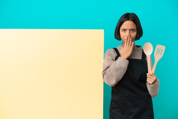 Zdjęcie młoda kobieta kucharz rasy mieszanej z dużym plakatem na białym tle na niebieskim tle obejmujące usta ręką