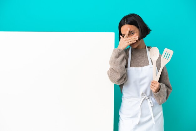 Młoda kobieta kucharz rasy mieszanej z dużym plakatem na białym tle na niebieskim tle zasłaniając oczy rękami i uśmiechając się