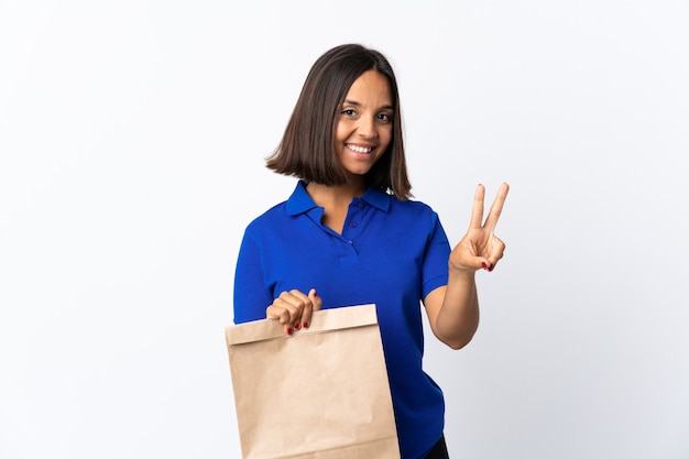 Młoda kobieta Łacińskiej trzyma torbę na zakupy spożywcze na białym tle uśmiecha się i pokazuje znak zwycięstwa