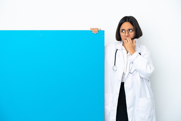 Młoda kobieta lekarz rasy mieszanej z dużą tabliczką na białym tle zdenerwowana i przestraszona przykładająca ręce do ust