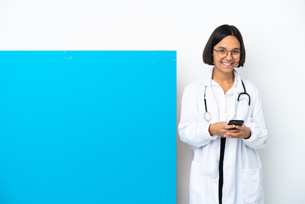 Młoda kobieta lekarza rasy mieszanej z dużą tabliczką na białym tle wysyłająca wiadomość za pomocą telefonu komórkowego