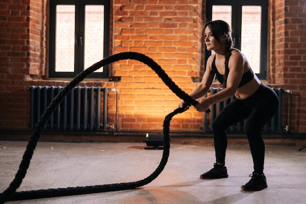 Młoda kobieta lekkoatletycznego fitness z silne piękne ciało w czarnej odzieży sportowej, ćwiczenia z linami o treningu treningowym. Muskularny trening kobiet rasy kaukaskiej w ciemnej siłowni.