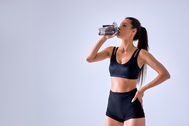 Młoda kobieta lekkoatletycznego picia świeżej wody po aktywnym treningu, trzyma butelkę wody, ubrana w czarną odzież sportową, na białym tle nad szarym betonowym murem. Koncepcja ludzie i znużenie. portret