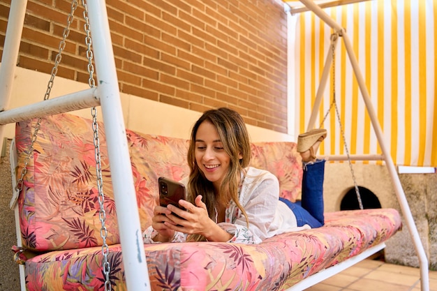 Młoda kobieta leżąca na huśtawce na podwórku korzystająca ze swojego smartfona rozmawiając z przyjaciółmi