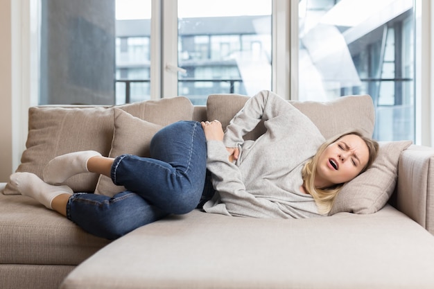 Młoda kobieta leżąca na kanapie w domu z silnym bólem brzucha