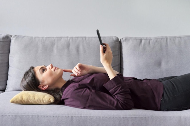 Młoda kobieta leżąca na kanapie za pomocą smartfona Kobieta marzycielsko uśmiecha się marzy pozytywne emocje