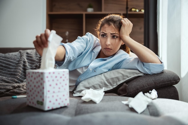 Zdjęcie młoda kobieta leży chora w domu na kanapie i bierze papierową chusteczkę do wydmuchiwania nosa.