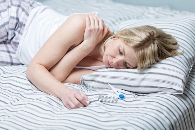 Młoda kobieta leży na łóżku z termometrem i tabletkami Blondynka w piżamie źle się czuje Choroby i alergie