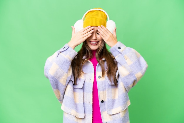 Młoda kobieta ma na sobie zimowe mufki na odizolowanym tle z kluczem chrominancji, zakrywającym oczy rękami