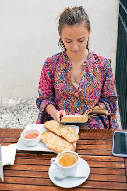 Młoda kobieta ma tost na śniadanie na tarasie baru i czyta książkę
