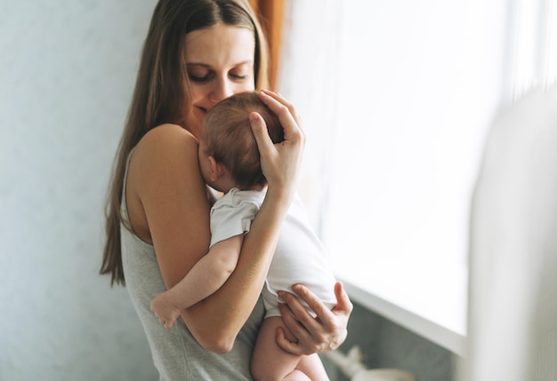 Młoda kobieta mama z długimi włosami trzymająca córeczkę na rękach w pobliżu okna w domu