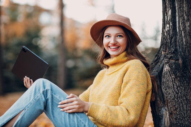 Młoda kobieta model siedzi z cyfrowym tabletem w jesiennym parku z żółtymi liśćmi klonu. Moda na jesień