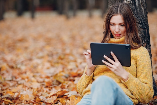 Młoda kobieta model siedzi z komputera typu tablet w jesiennym parku z żółtymi liśćmi klonu. Moda na jesień