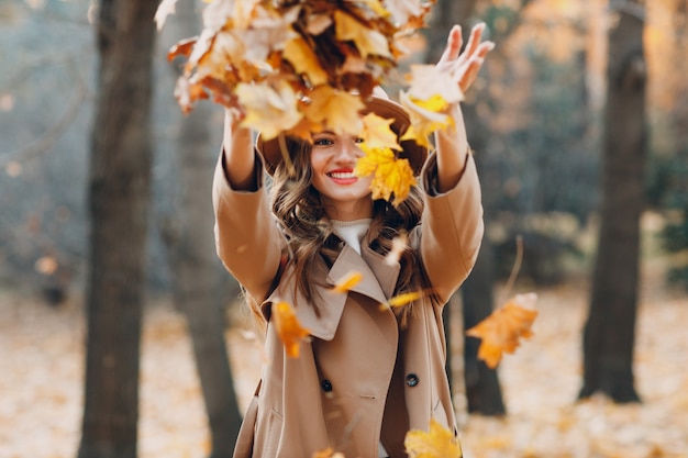Młoda kobieta model w jesiennym parku rzucać żółte liście klonu liści. Moda na jesień