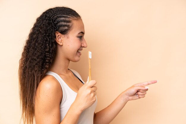 Młoda kobieta myjąca zęby odizolowana na beżowym tle, wskazująca na bok, aby zaprezentować produkt