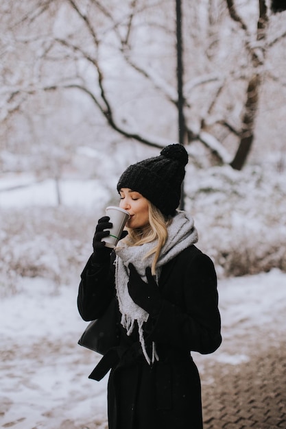 Młoda kobieta n ciepłe ubrania, ciesząc się w śniegu z filiżanką kawy na wynos