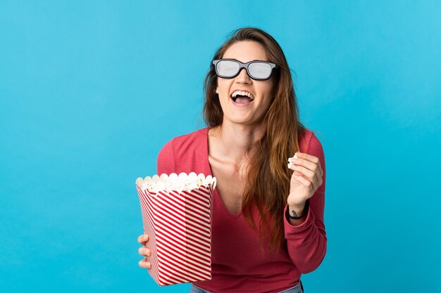 Młoda kobieta na białym tle na niebieskiej ścianie z okularami 3d i trzymając duże wiadro popcorns, wskazując przód