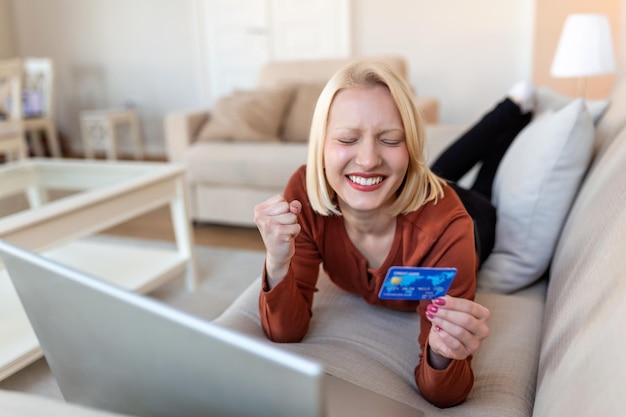 Młoda kobieta na kanapie robi zakupy online z kartą debetową Piękna dziewczyna używa laptopa do zakupów online w domu