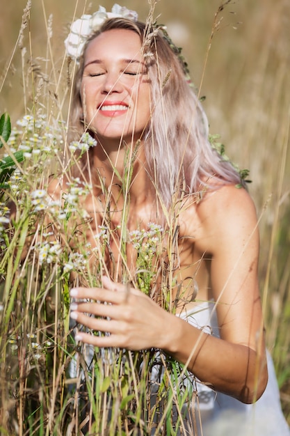 Młoda kobieta na polu rozkoszuje się aromatem ziół. Uśmiechnięta blondynka w białej sukni i wianku. Pionowy.