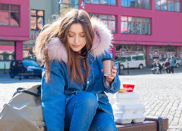 Zdjęcie młoda kobieta na ulicy w płaszczu z pudełkami na lunch i dostawą zabiera kawę i jedzenie.