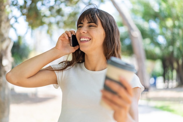 Młoda kobieta na zewnątrz za pomocą telefonu komórkowego i trzymając kawę z radosnym wyrazem twarzy