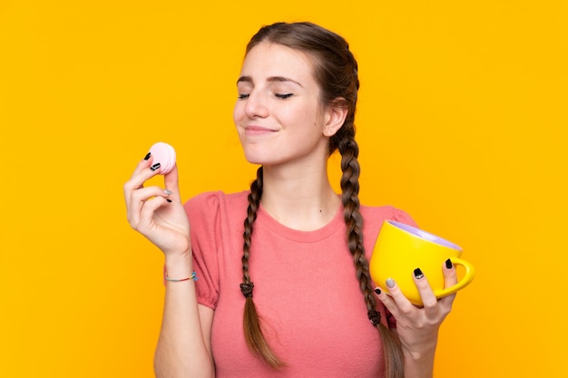 Młoda kobieta nad odosobnioną kolor żółty ścianą trzyma kolorowych Francuskich macarons i filiżankę mleka