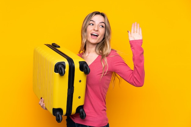 Młoda kobieta nad odosobnioną kolor żółty ścianą w wakacje z podróży walizką i salutować