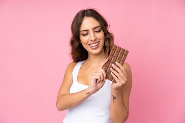 Młoda kobieta nad różową ścianą bierze czekoladową pastylkę i szczęśliwa