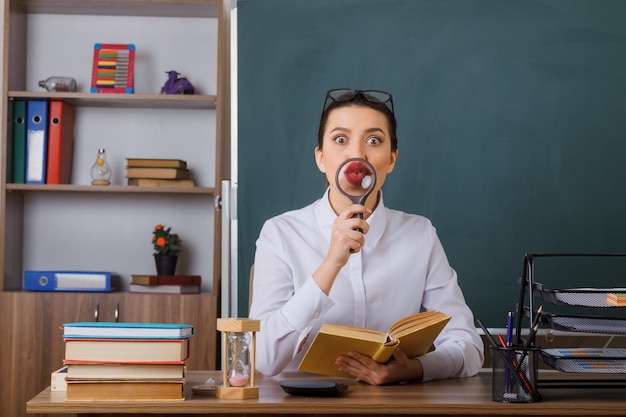 Młoda Kobieta Nauczyciel W Okularach Siedzi Przy ławce Szkolnej Z Książką Przed Tablicą W Klasie, Trzymając Szkło Powiększające Przed Ustami