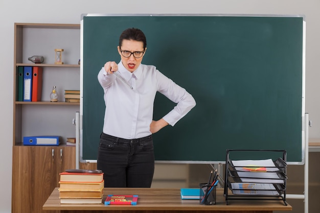 Młoda kobieta nauczycielka w okularach wyjaśniająca lekcję wskazującą ze wskaźnikiem na kamerę będąc niezadowolony stojąc przy ławce szkolnej przed tablicą w klasie