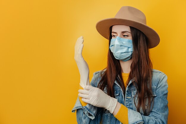 Młoda kobieta nosi ochronną maskę medyczną, aby nie zarazić innych ludzi, zakłada rękawiczki medyczne, przygotowując się do niebezpiecznej podróży podczas wybuchu epidemii koronawirusa, na białym tle na żółtym tle