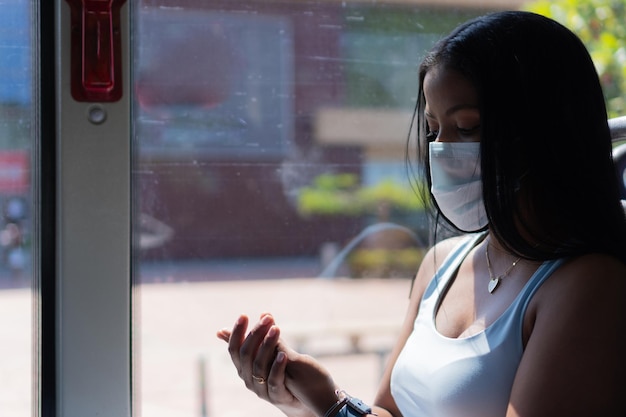 Młoda kobieta nosząca maskę rozpylającą środek dezynfekujący pod ręką podczas jazdy autobusem podczas pandemii