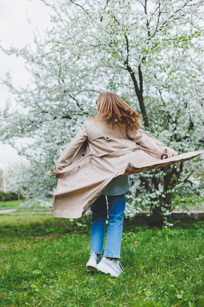 Młoda kobieta o blond włosach cieszy się kwitnącym wiosennym ogrodem Przerwa wiosenna w podróży Modny styl Kobieta w okularach przeciwsłonecznych i beżowym trenczu biegnie przez kwitnący park Ostrość na selekcji