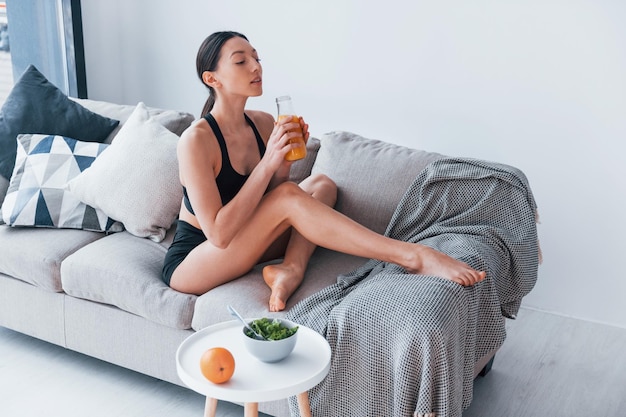 Młoda kobieta o szczupłej sylwetce w odzieży sportowej siedzi na kanapie i je zdrową dietę w domu w domu