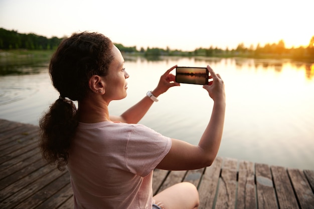 Zdjęcie młoda kobieta odpoczywa na molo i fotografuje piękny zachód słońca nad jeziorem w letni wieczór