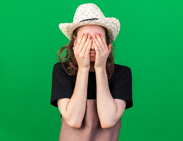 Młoda kobieta ogrodniczka w kapeluszu ogrodniczym z zakrytymi oczami rękami odizolowanymi na zielonej ścianie