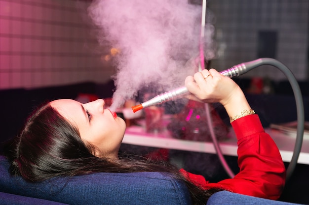 Młoda kobieta pali fajkę wodną lub fajkę wodną w klubie lub pali dymek