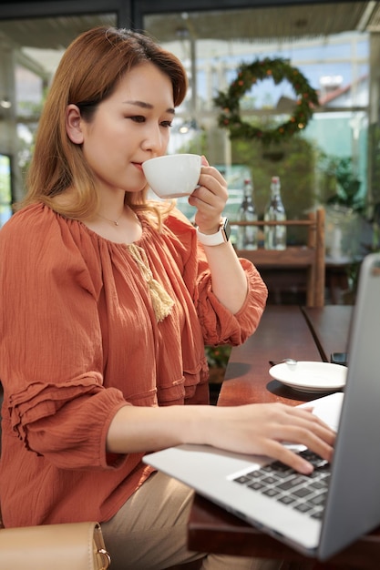 Młoda kobieta pije filiżankę porannej kawy i pracuje na laptopie w kawiarni