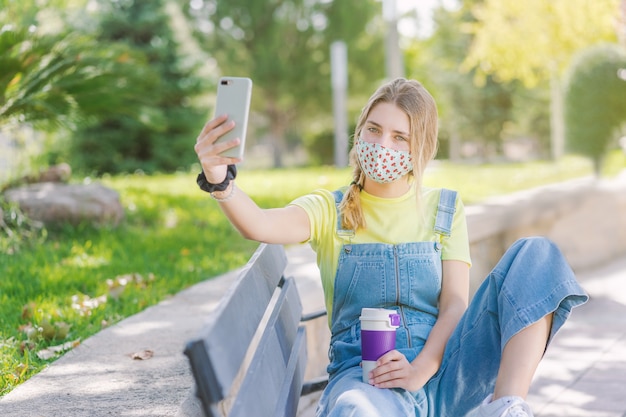 Młoda kobieta pije kawę i robi selfie w parku podczas pandemii