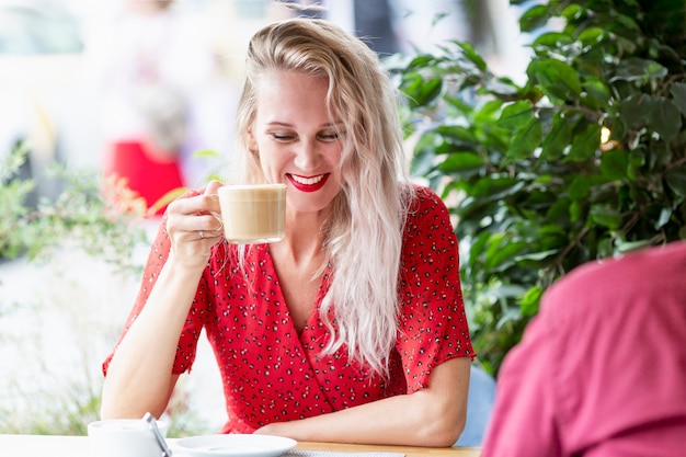 Młoda Kobieta Pije Kawę I Się śmieje. Piękna Blondynka Z Długimi Włosami W Czerwonej Sukience Na Tarasie Letniej Kawiarni.